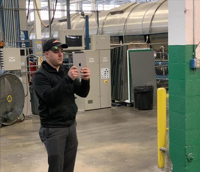 A man wearing a SERVPRO hat holding an iPad standing in a room near industrial machinery